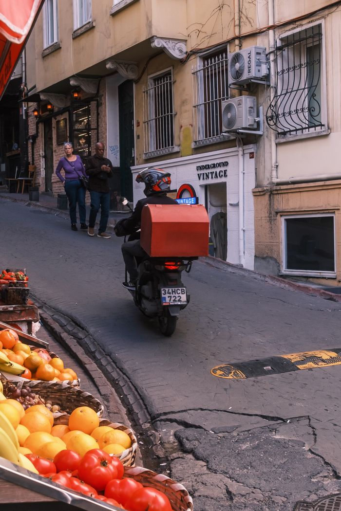 Street view of a scooter delivery on a vibrant urban road with fruit stalls.